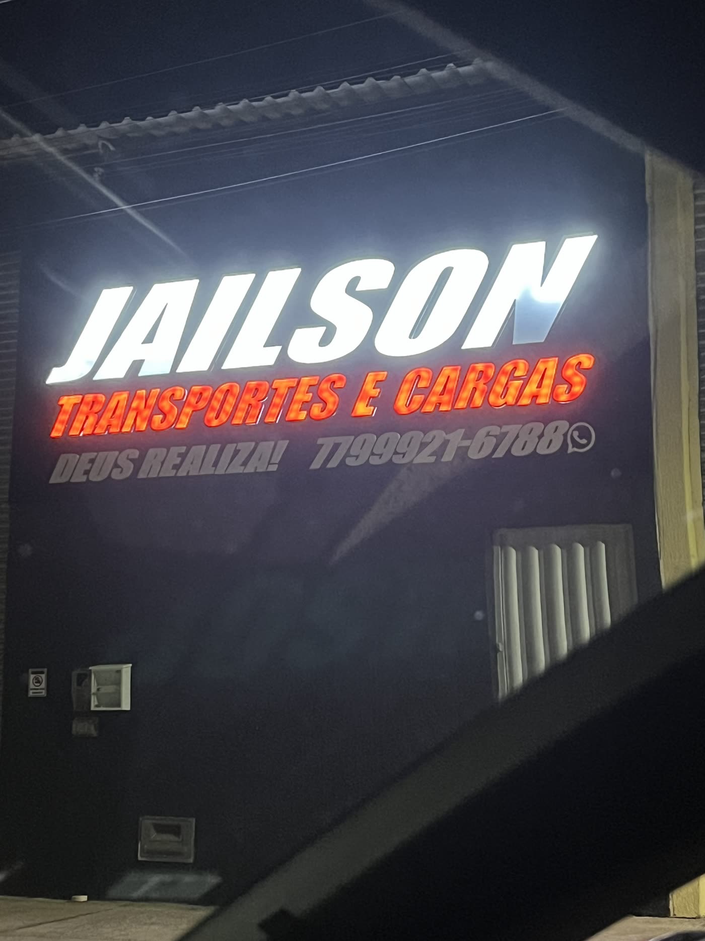 Letreiro Jailson Transportes e Cargas aceso na fachada do galpão, à noite